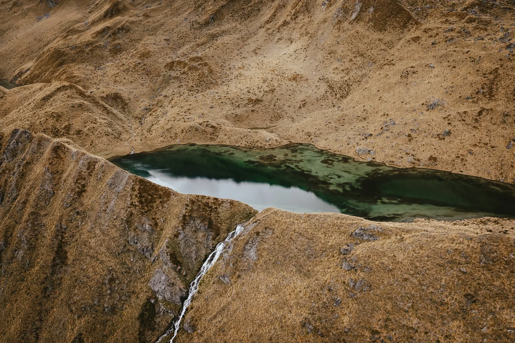 An aerial view of a small emerald alpine tarn set into golden tussock slopes near Wanaka.