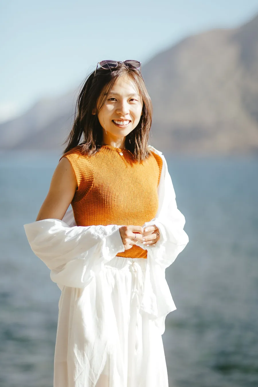 Arabella smiling by a lake in an orange top and white jacket, mountains behind her.