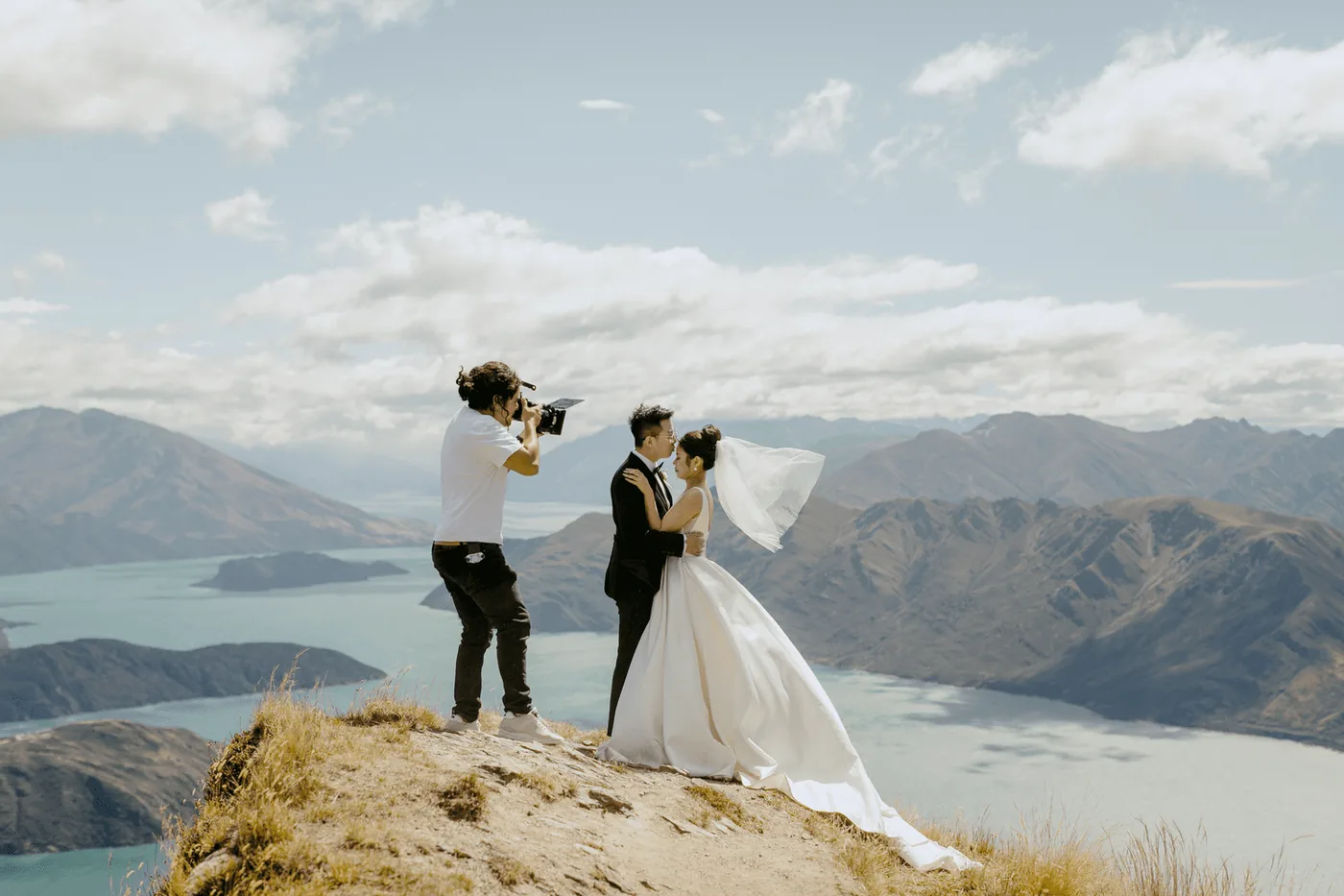 A videographer filming an eloping couple embracing on a ridge above a lake in Queenstown.