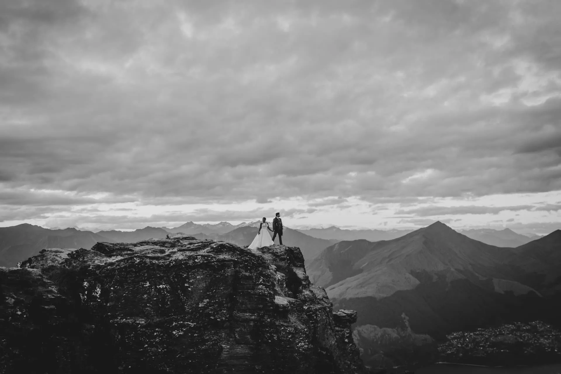 Black and white photo of a couple on a Cecil Peak rock outcrop with mountain ranges and cloud behind.