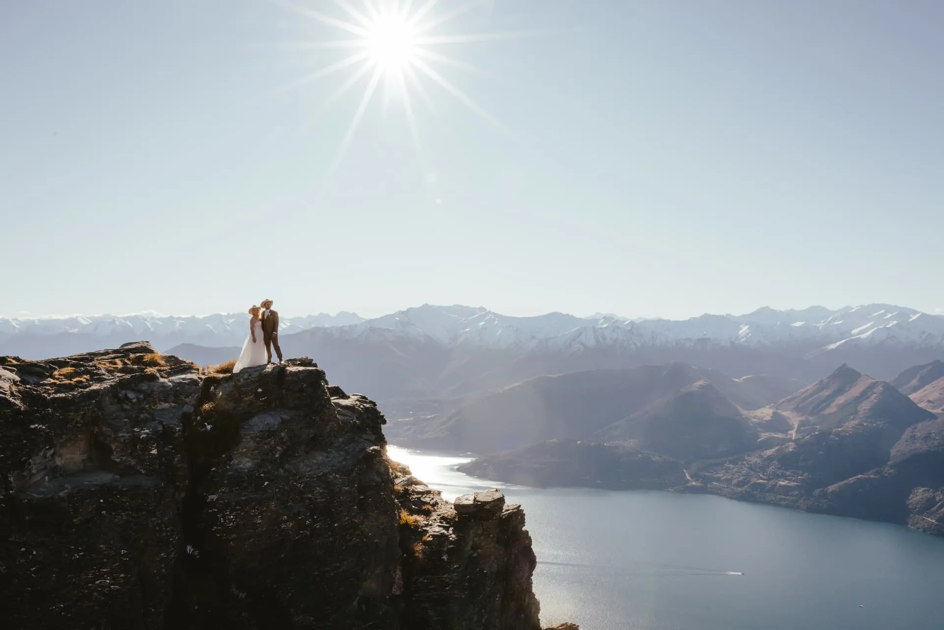 Couple standing on a Cecil Peak cliff top with the snowy Southern Alps stretching behind them.