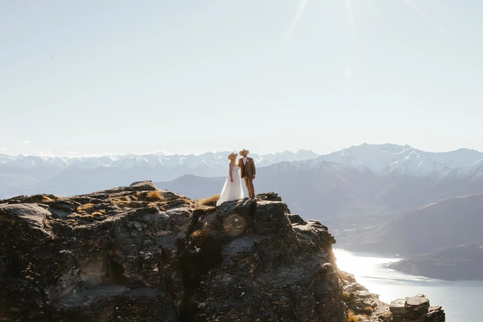 Couple embracing beside their helicopter on golden tussock on Cecil Peak, a snow-covered mountain behind.