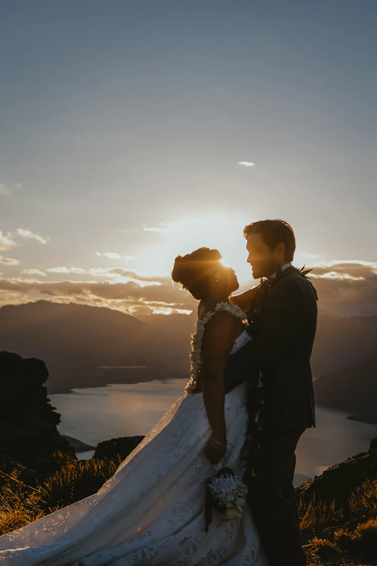 Bride and groom close together at sunset on Cecil Peak, Lake Wakatipu and the mountains behind them.