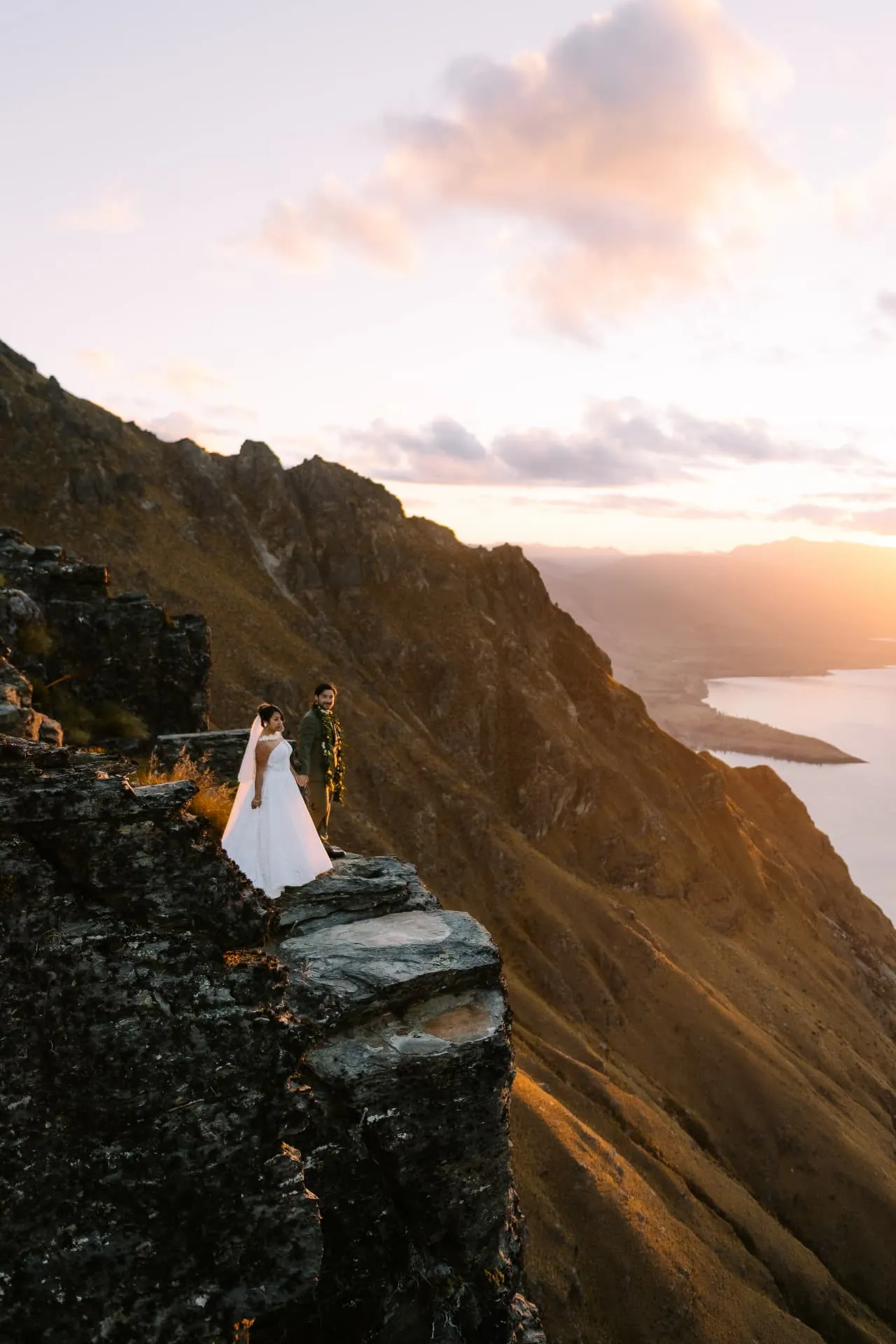 Vertical view of a couple standing on the Cecil Peak Ledge cliff above Lake Wakatipu at sunset.