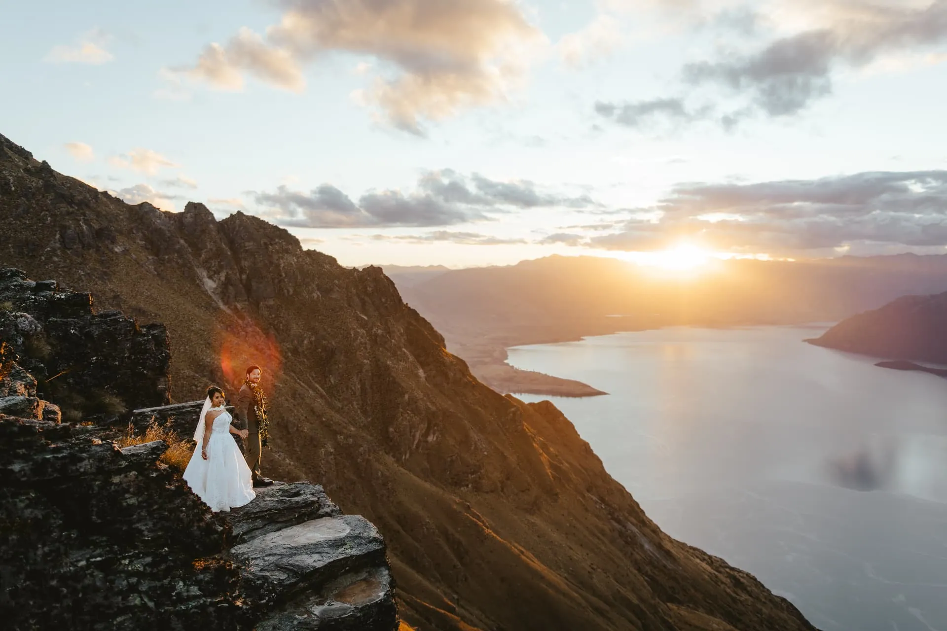 Bride and groom on the cliff edge of the Cecil Peak Ledge, a sheer face dropping to Lake Wakatipu at sunset.