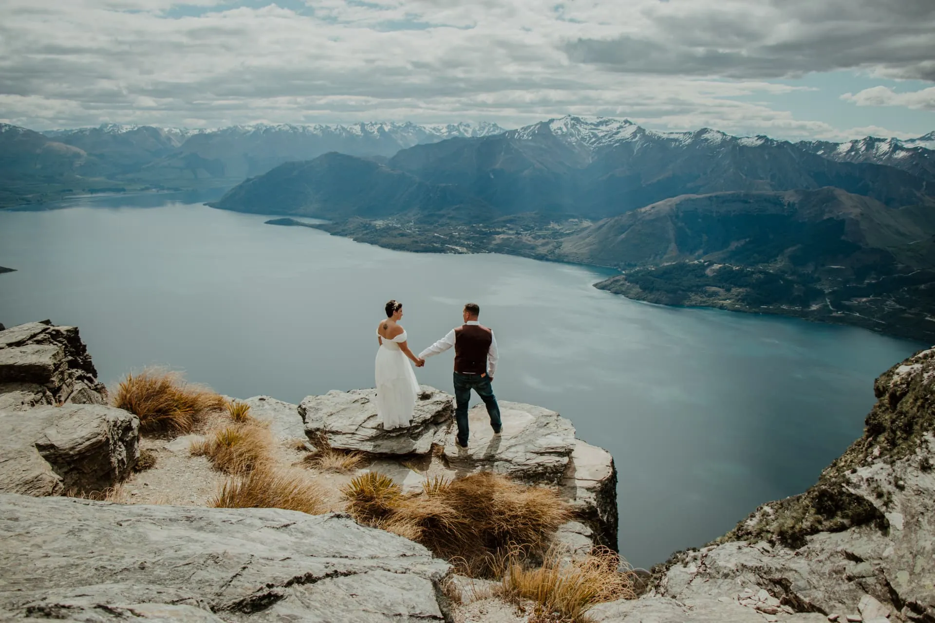 Couple holding hands at the cliff edge of the Cecil Peak Ledge among golden tussock, lake below.