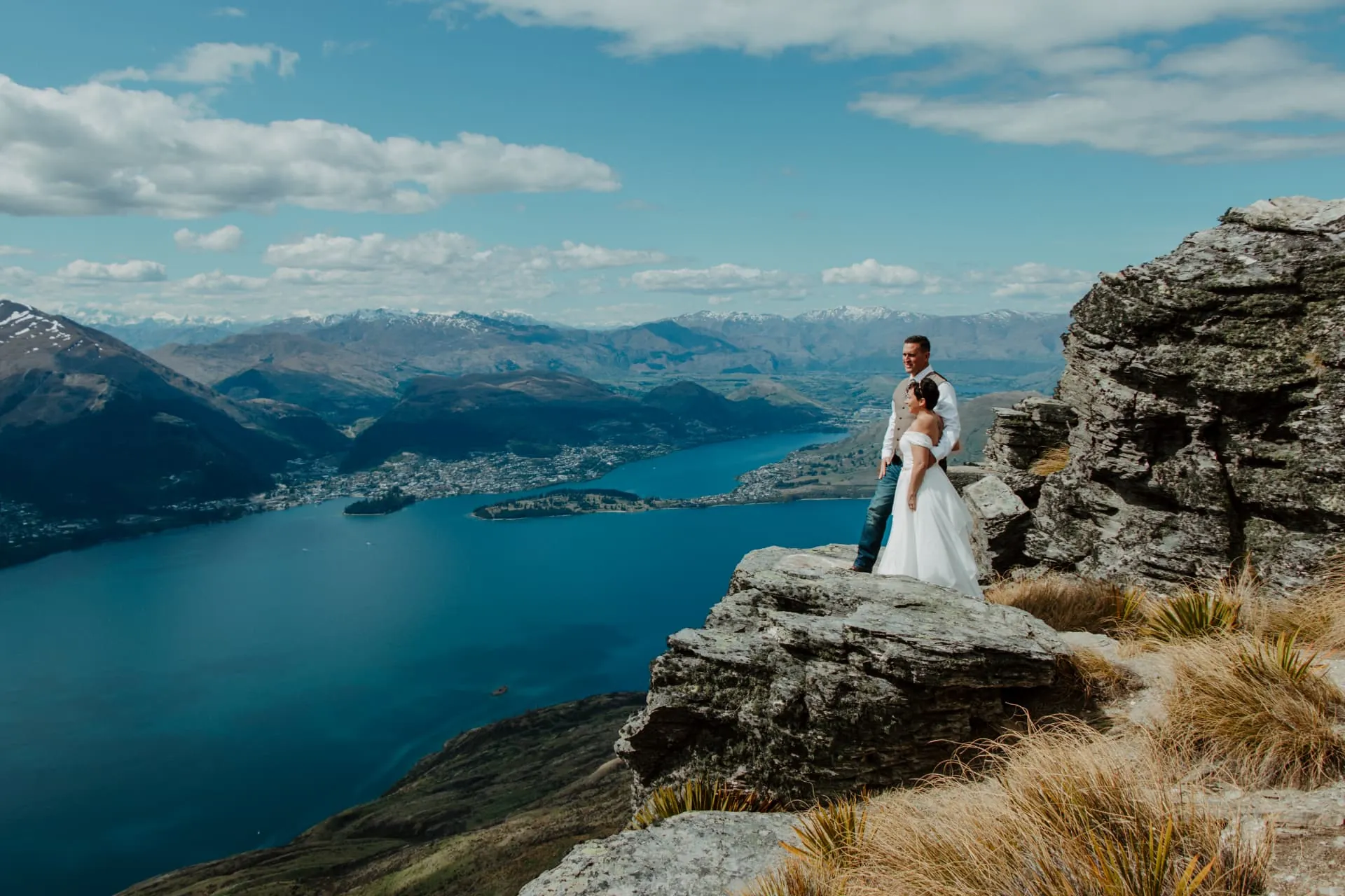 Bride and groom on a rock outcrop on the Cecil Peak Ledge above a deep blue Lake Wakatipu.