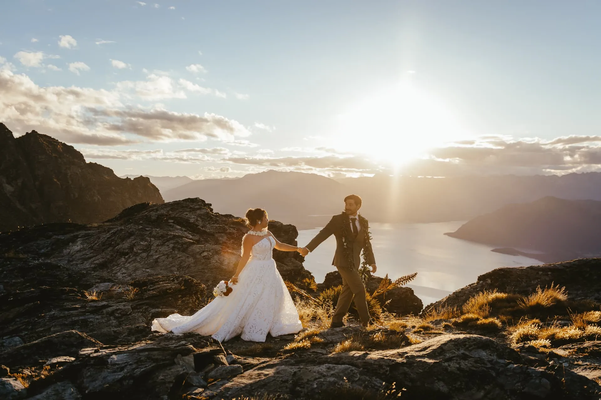 Couple walking across schist rock and golden tussock on the Cecil Peak Ledge at golden hour above Lake Wakatipu.