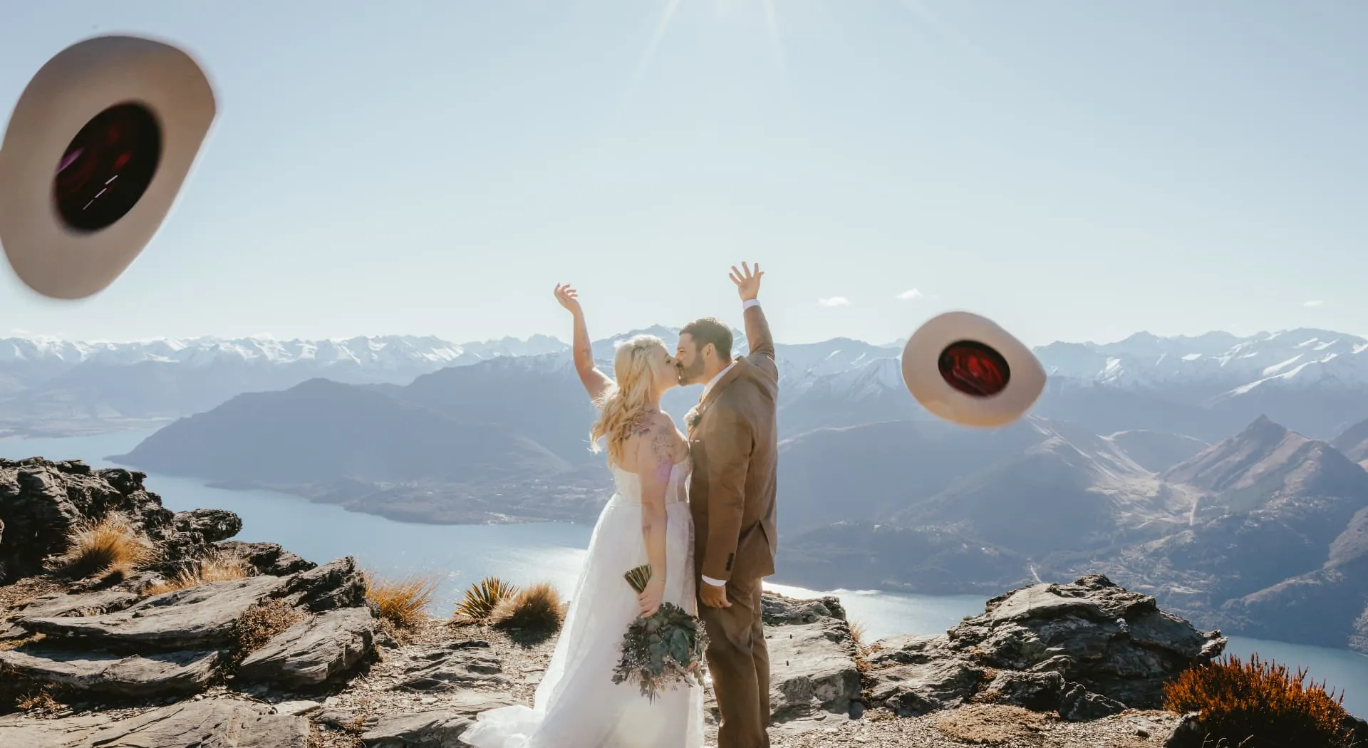Newlyweds tossing their hats on the Cecil Peak Ledge, Lake Wakatipu and the Queenstown mountains behind.