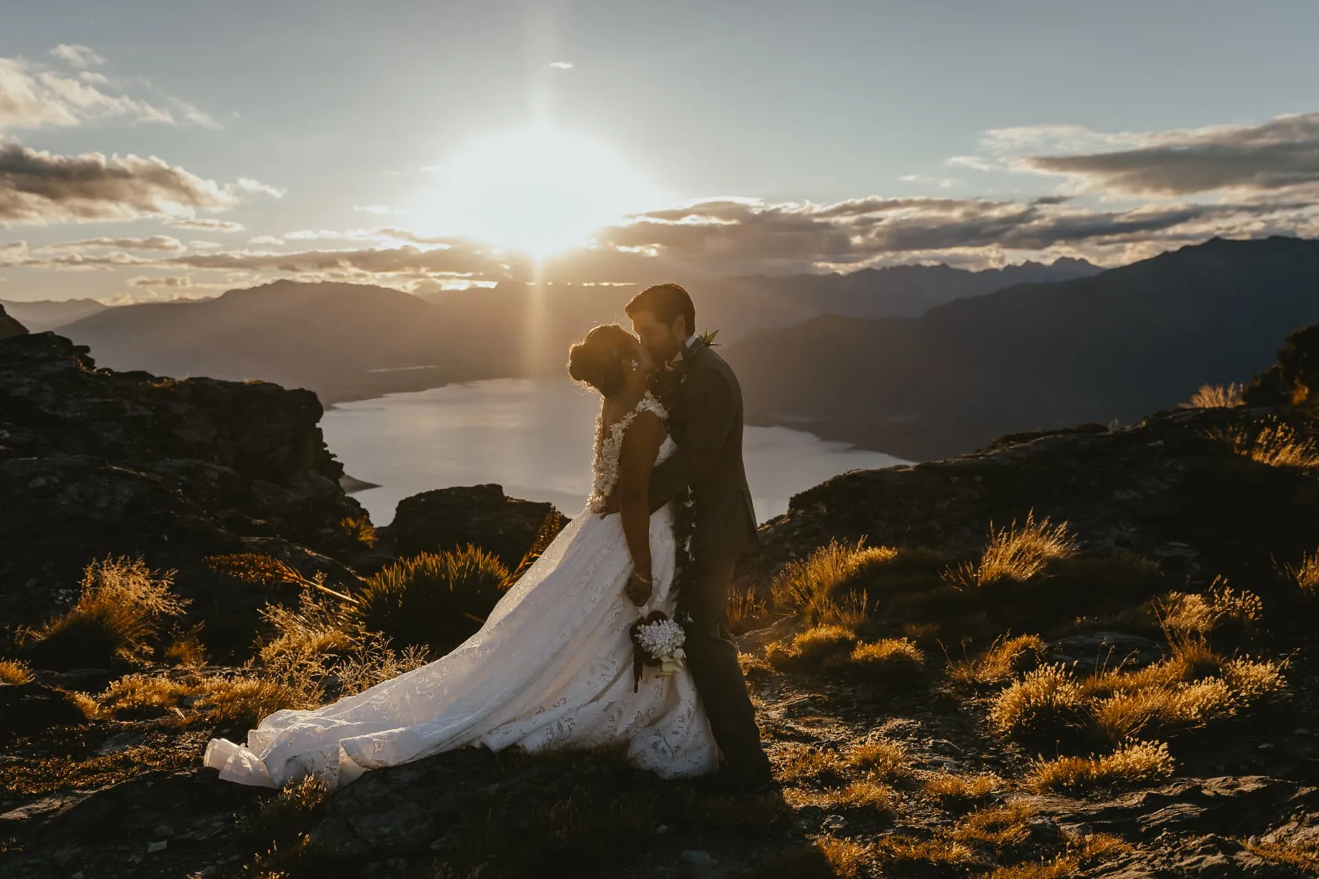 Bride and groom kissing on the Cecil Peak Ledge at sunset, the train of her dress across the tussock, Lake Wakatipu behind.