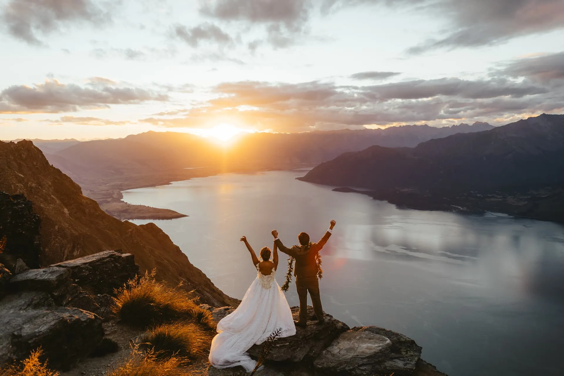 Newlyweds with arms raised on the Cecil Peak Ledge as the sun sets over Lake Wakatipu, Queenstown.