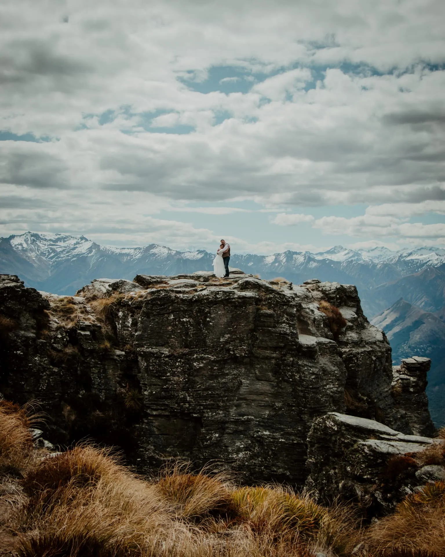 Couple standing on a tall rock pinnacle at Cecil Peak with the snowy Southern Alps behind.