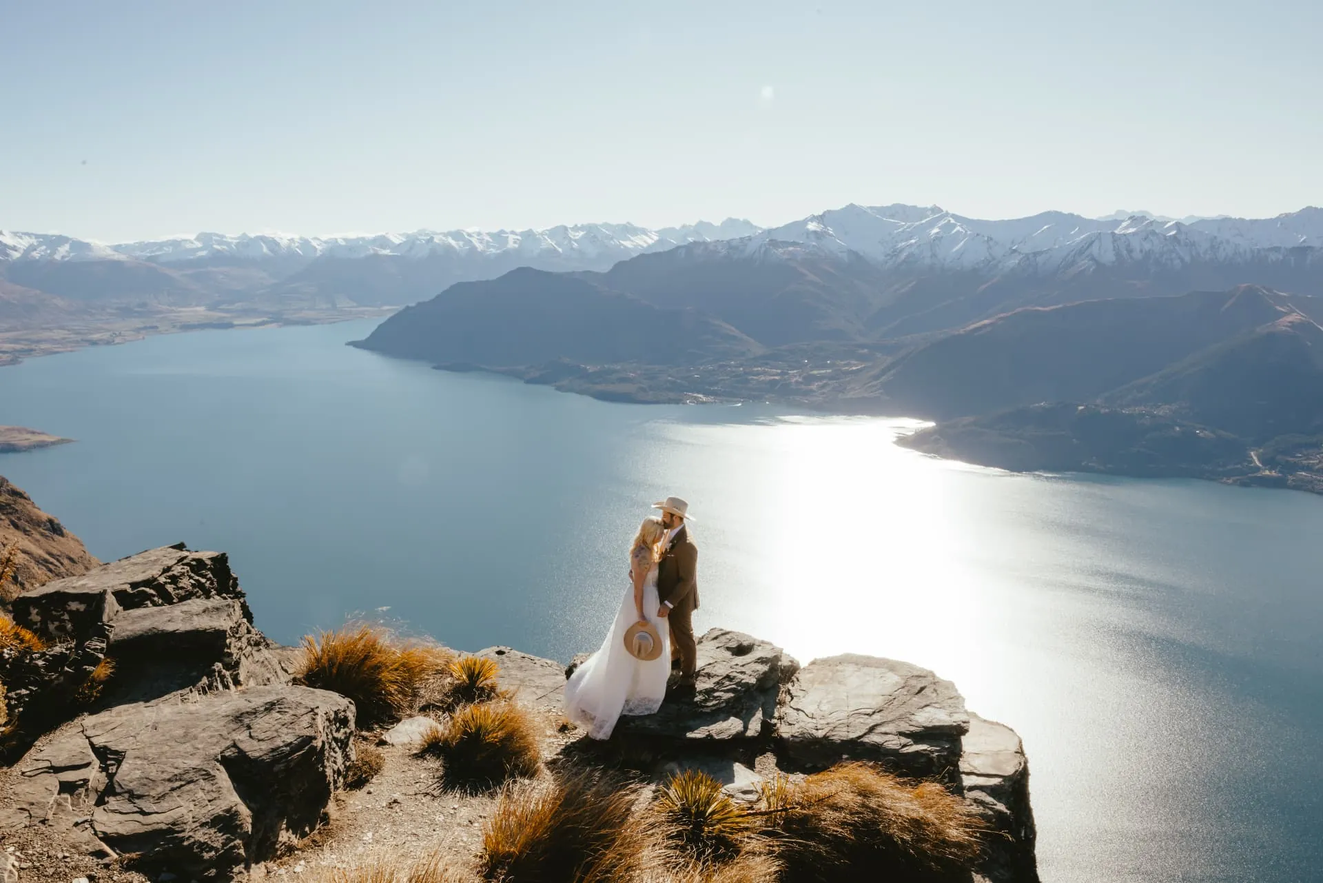 Couple on the Cecil Peak Ledge in spring, Lake Wakatipu shining below and snow-capped mountains beyond.