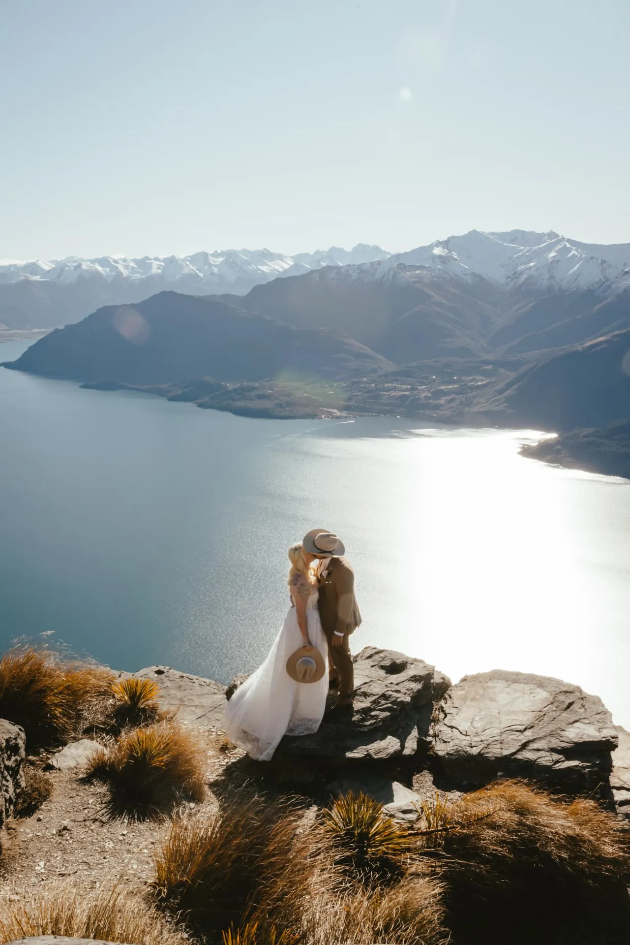 Couple embracing on the Cecil Peak Ledge in spring sunshine, the length of Lake Wakatipu behind them.