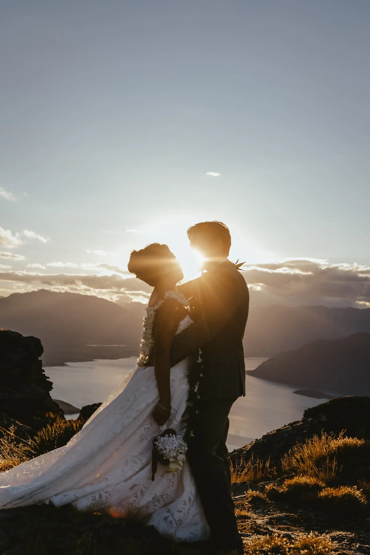Sun flaring between a couple embracing on the Cecil Peak Ledge, a Queenstown heli elopement at sunset.