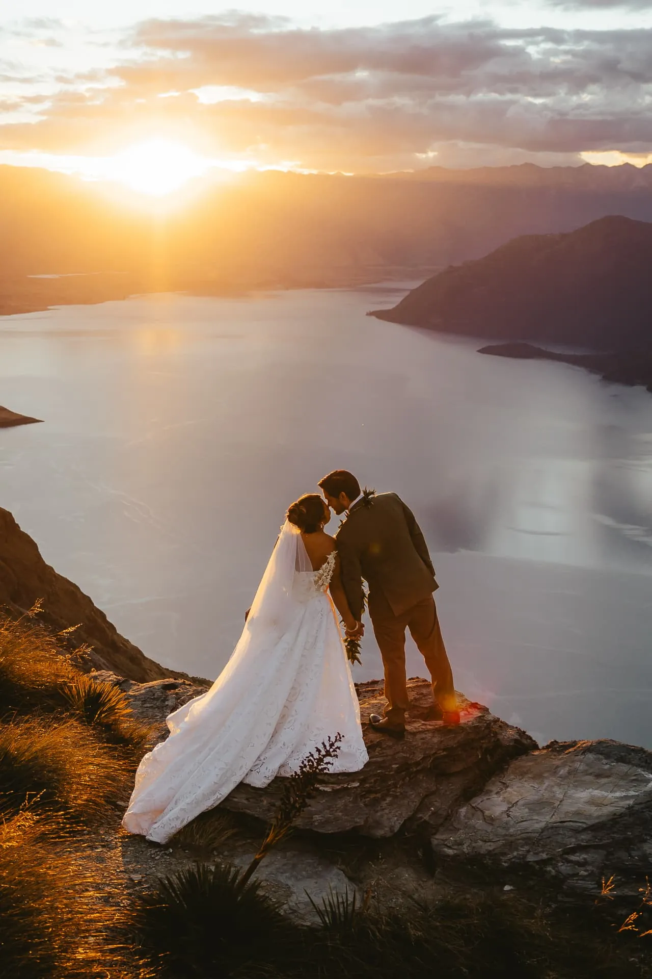 Bride and groom kissing on the Cecil Peak Ledge with the sun setting over Lake Wakatipu.