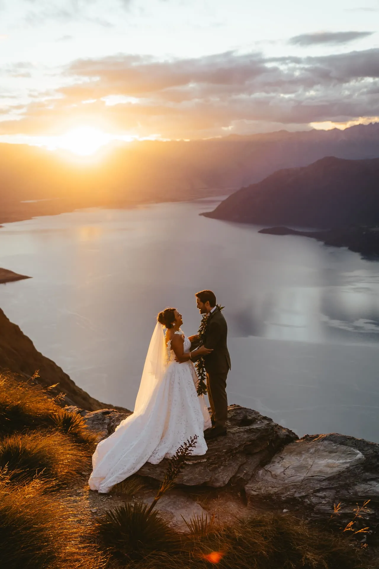 Bride with her veil and groom facing each other at sunset on the Cecil Peak Ledge, Lake Wakatipu below.
