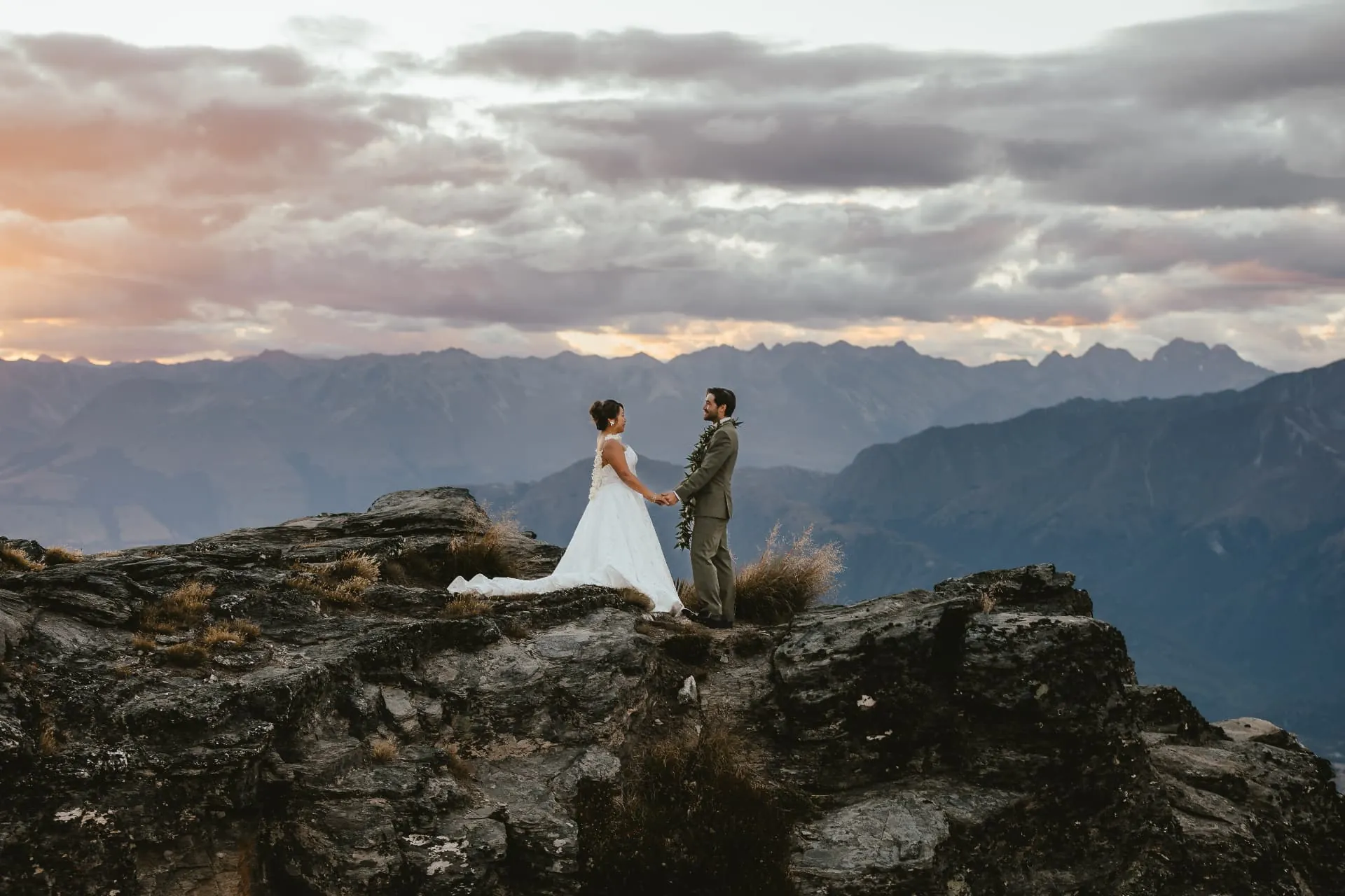 Couple holding hands during their wedding ceremony on the rocky Cecil Peak Ledge, mountains and cloud behind them, Queenstown.
