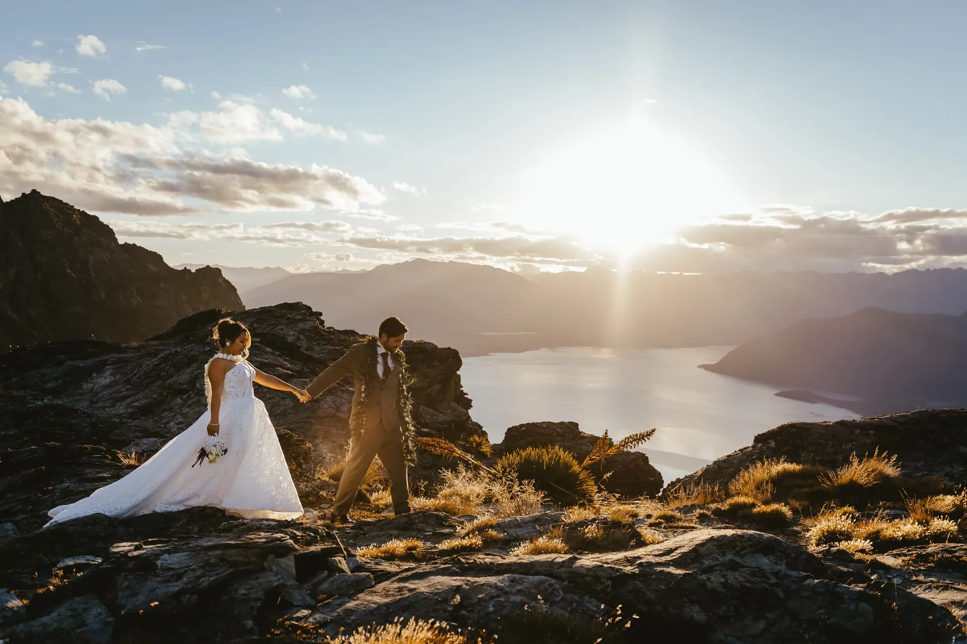 Bride and groom walking hand in hand on the Cecil Peak Ledge at sunset, Lake Wakatipu below, Queenstown.