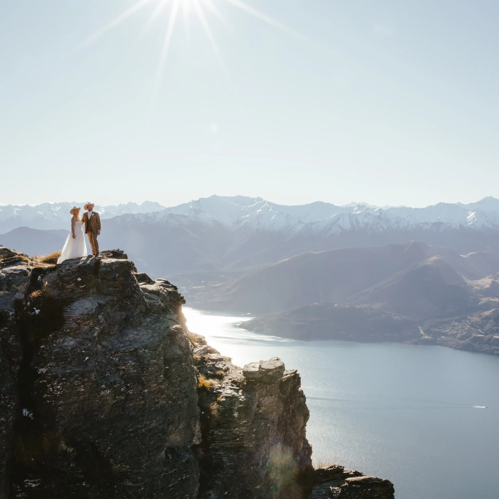 Couple on a rock pinnacle on Cecil Peak in spring, the lake winding away beneath snowy mountains.