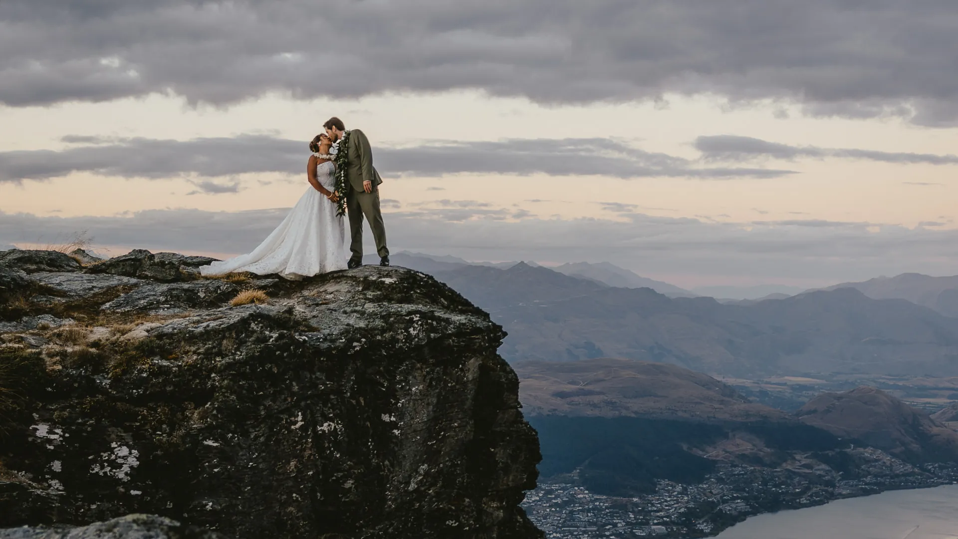 Couple standing on a rocky outcrop on Cecil Peak above Queenstown, with the town and lake visible below.