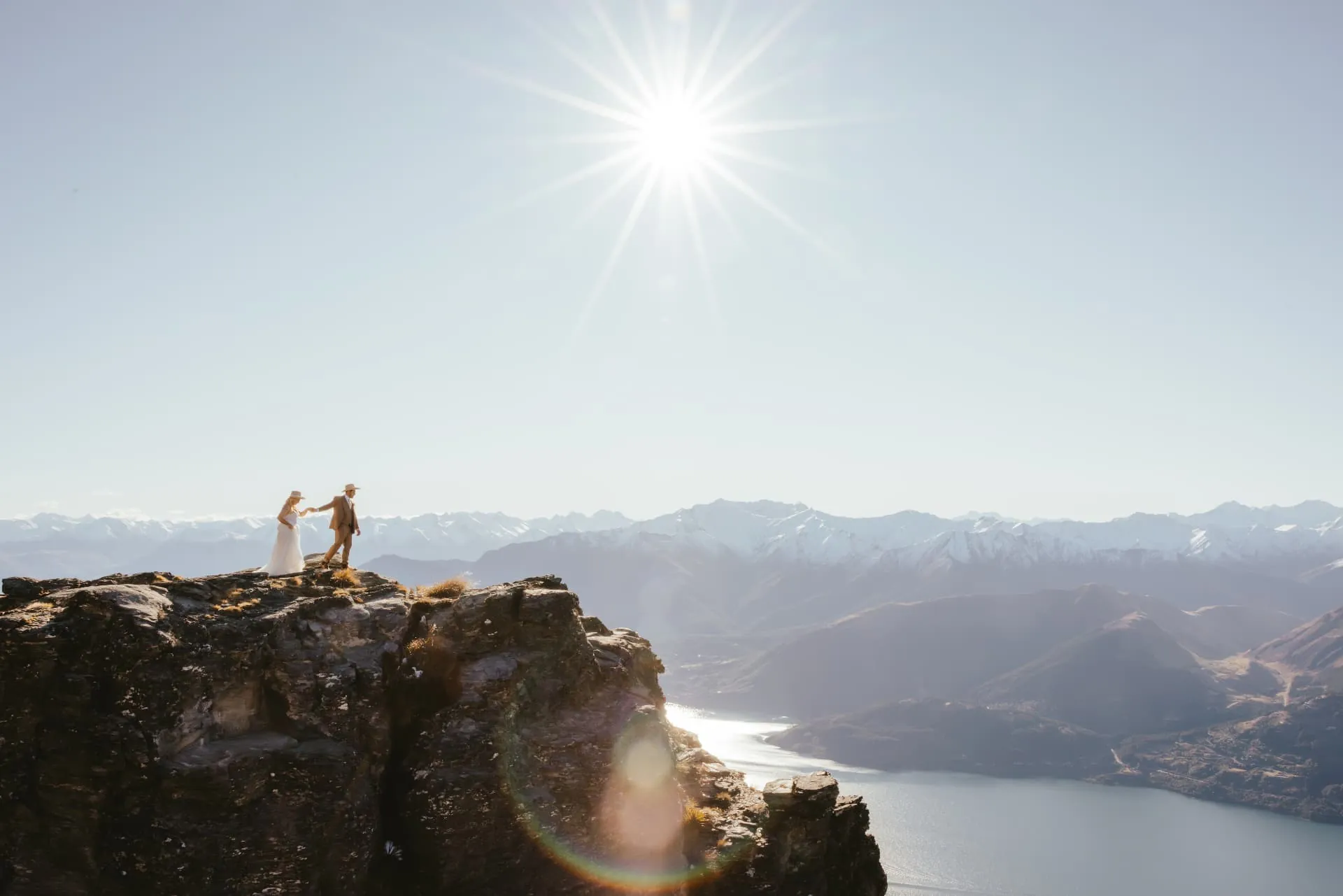 Couple on top of a rock outcrop on Cecil Peak with a spring sun flare and Lake Wakatipu below.
