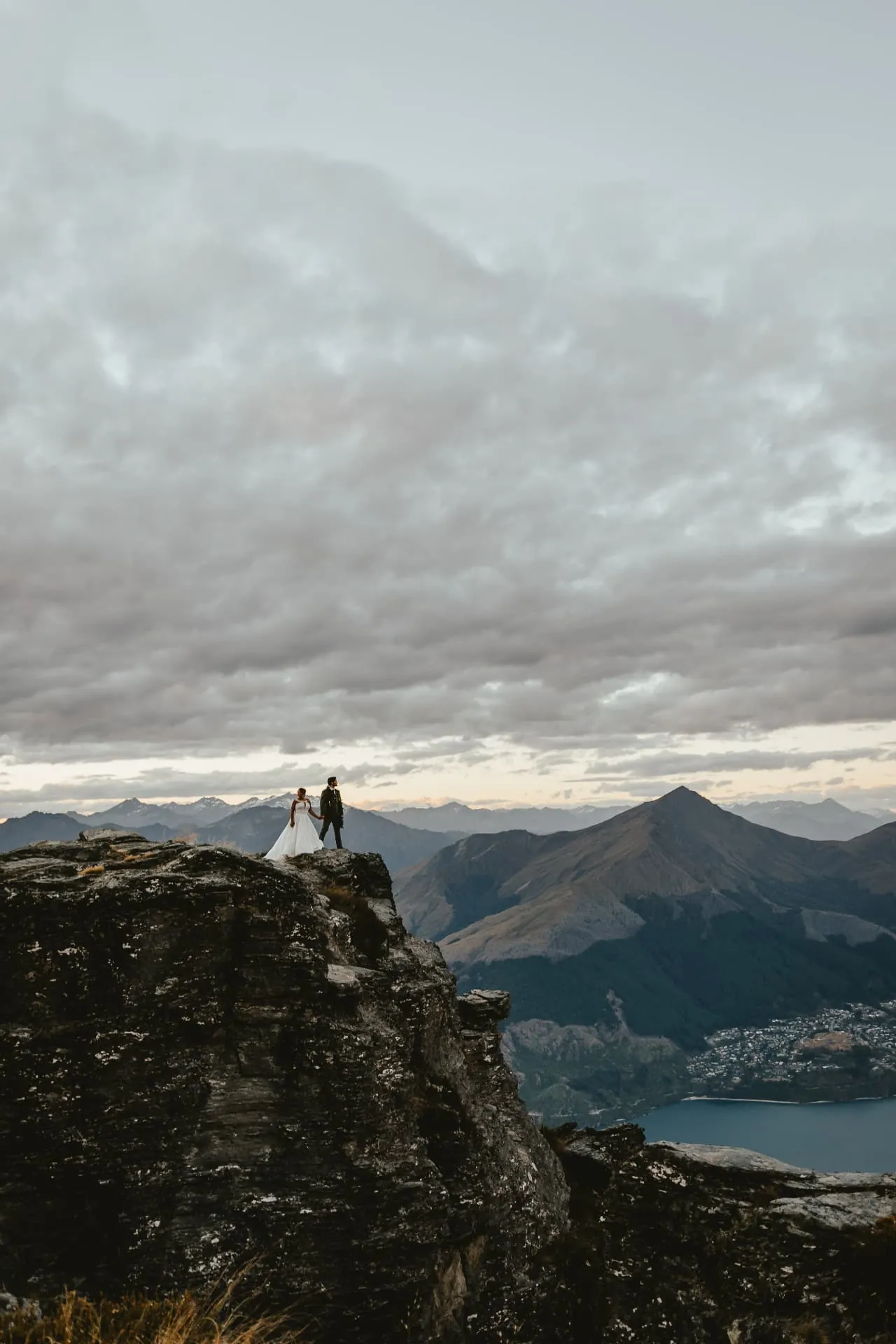 Couple tiny on top of a rock pinnacle at Cecil Peak beneath a dramatic cloudy sky, Queenstown.