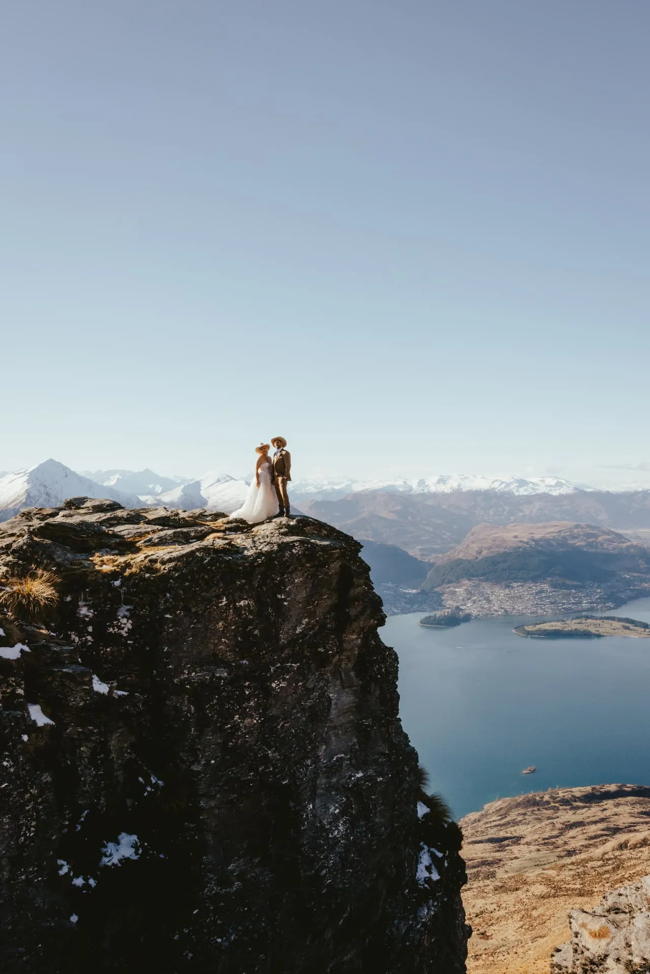 Couple small on top of a rock tower on Cecil Peak, Queenstown visible across Lake Wakatipu.