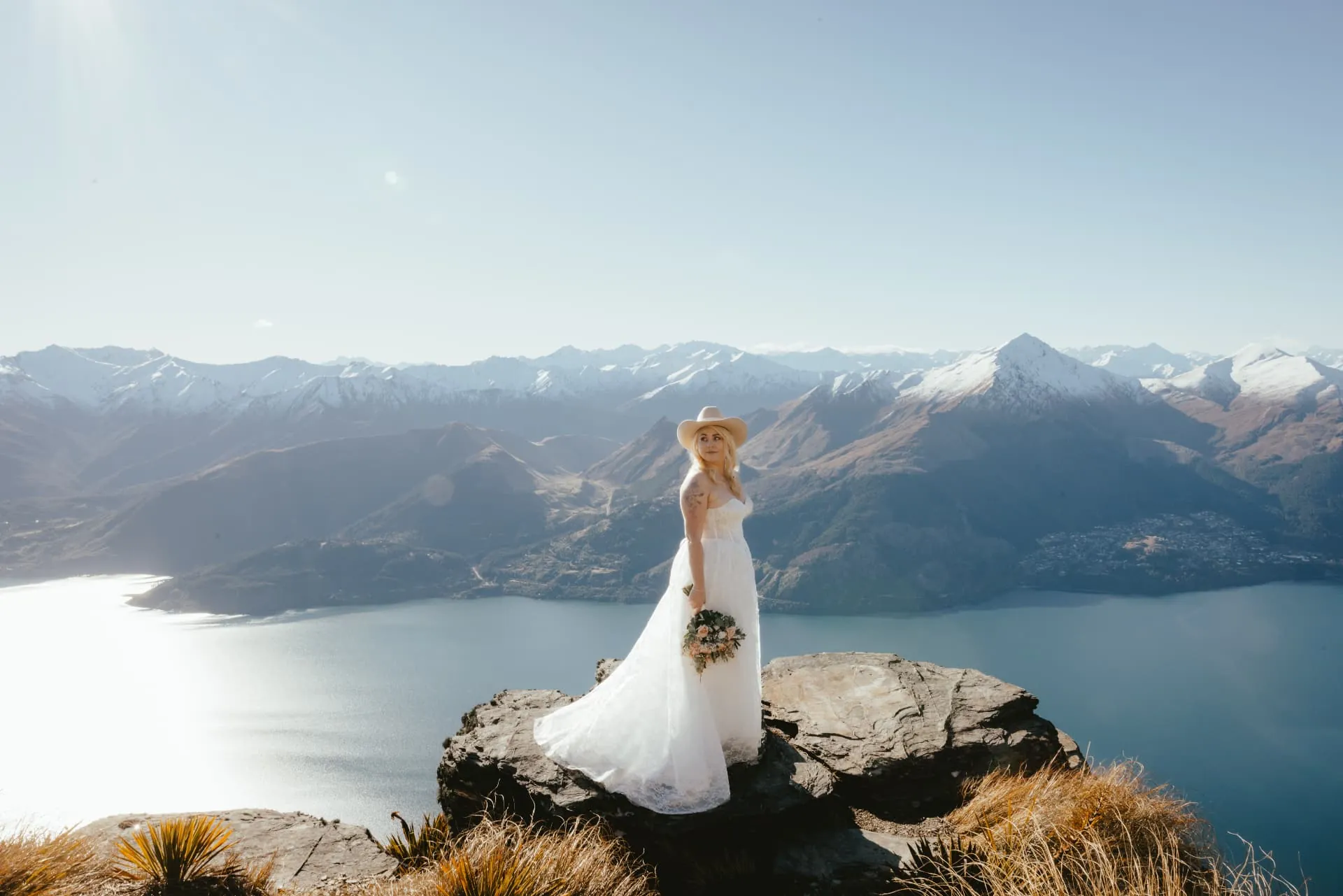 Spring bride in a hat on a rock above Lake Wakatipu, Queenstown and snow-capped ranges across the water from Cecil Peak.