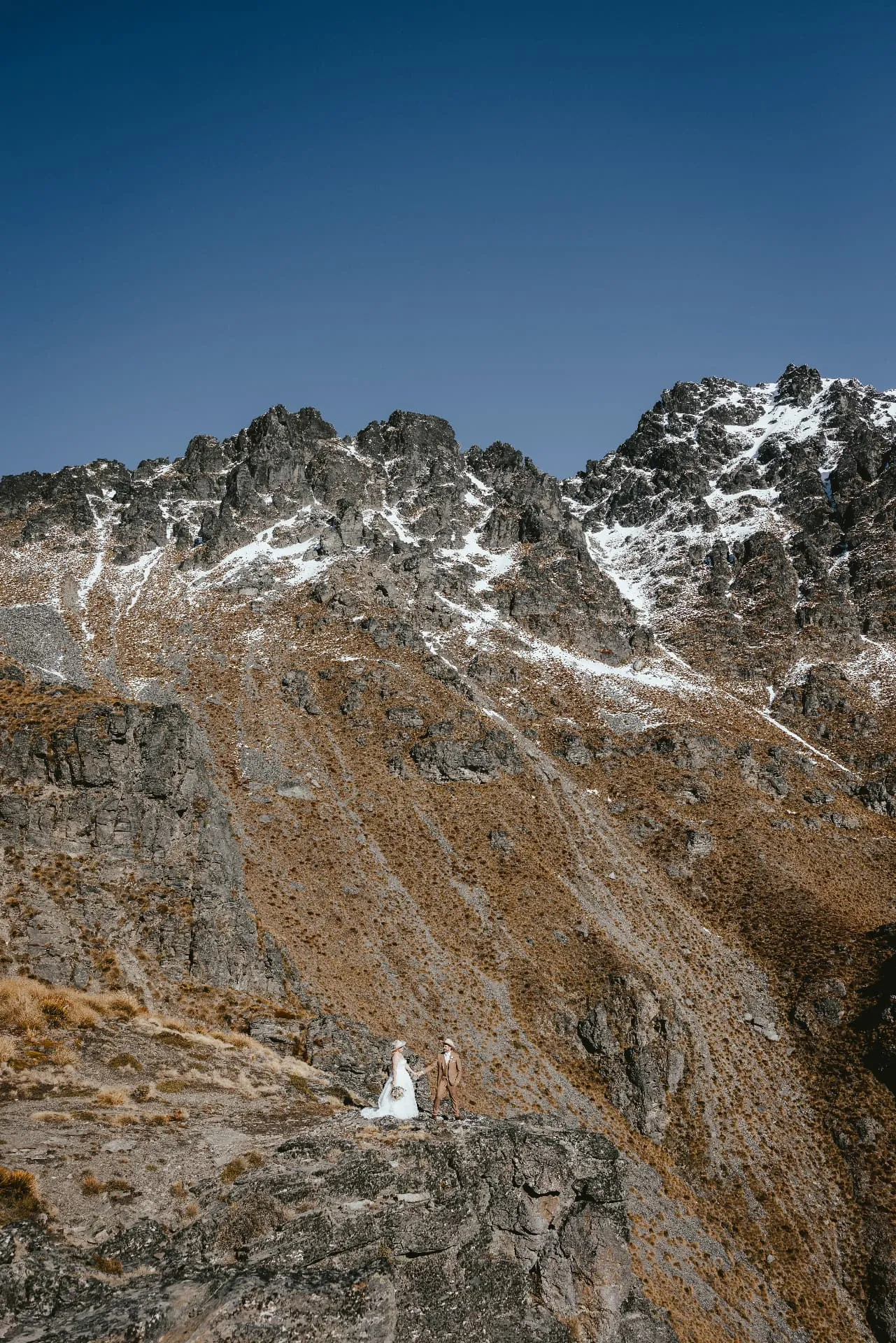 Spring snow on the craggy summit of Cecil Peak, with a wedding couple small on the tussock slope below.