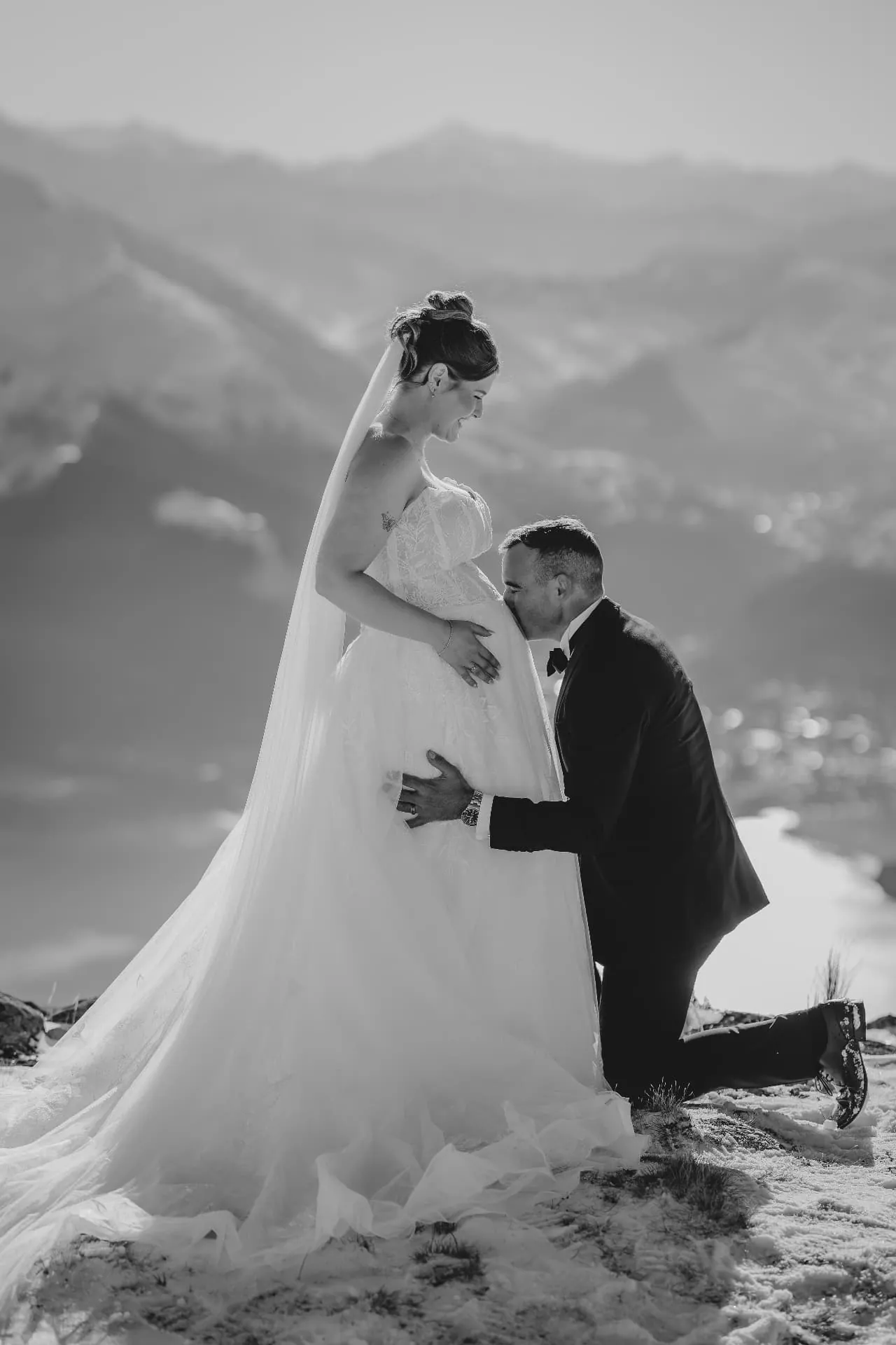 Black and white photo of a groom kissing his expecting bride’s baby bump during their Cecil Peak winter elopement.