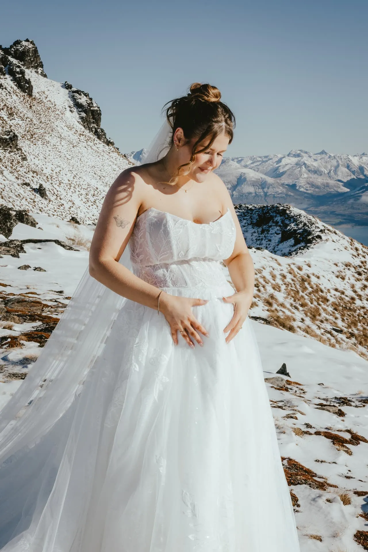 Bride smiling down in the snow on the Cecil Peak Spur, snowy ridge and mountains behind her.