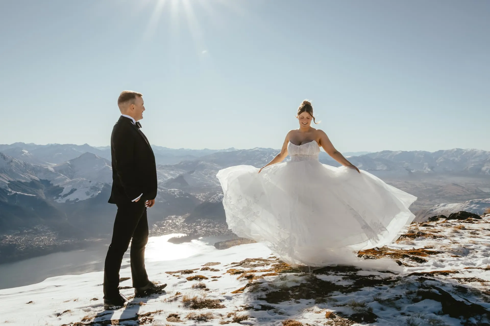 Bride twirling her dress on the snowy Spur at Cecil Peak, groom watching, Lake Wakatipu below.