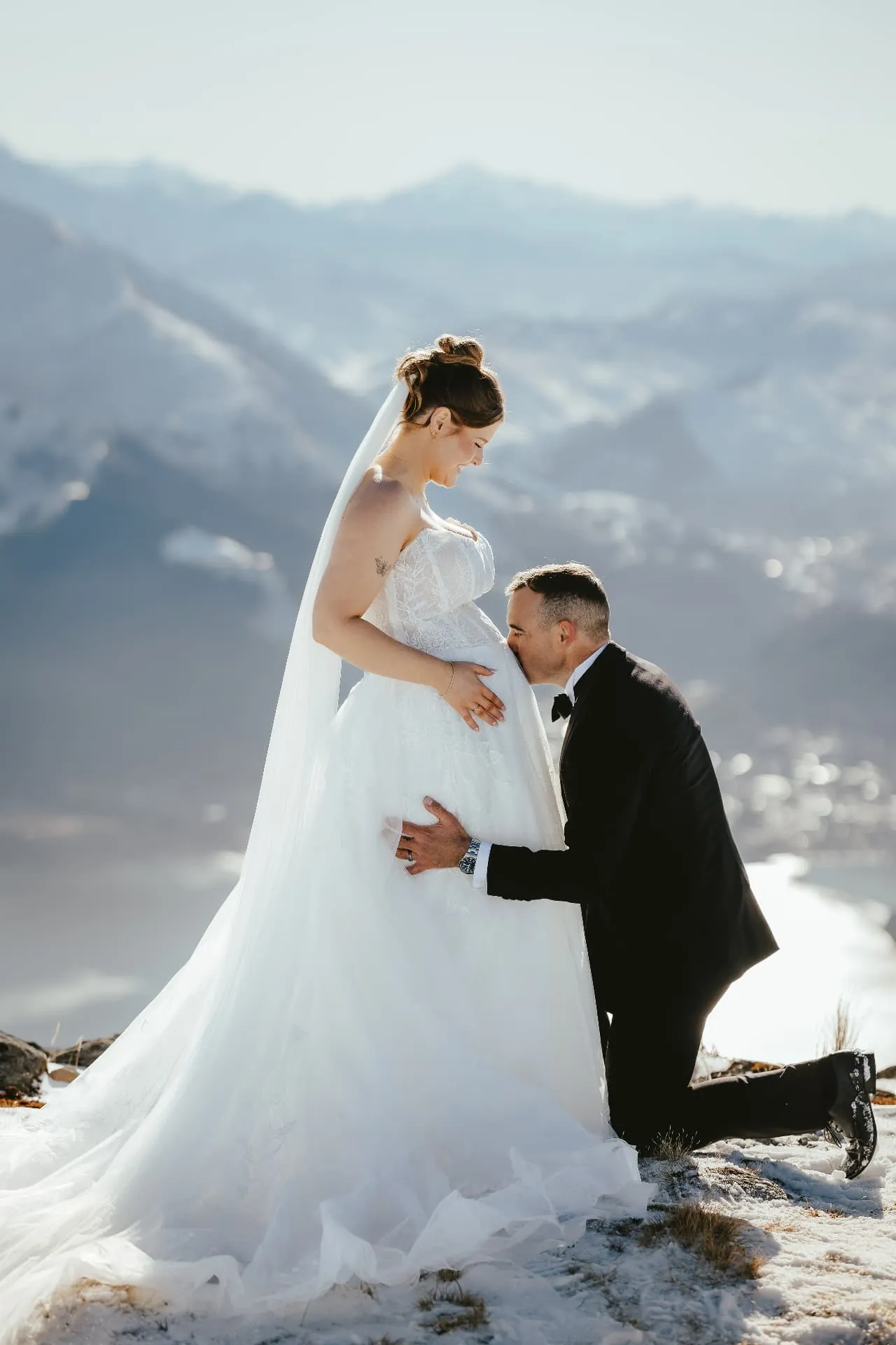 Groom kneeling to kiss his pregnant bride’s belly in the snow on the Cecil Peak Spur, Queenstown, mountains behind.