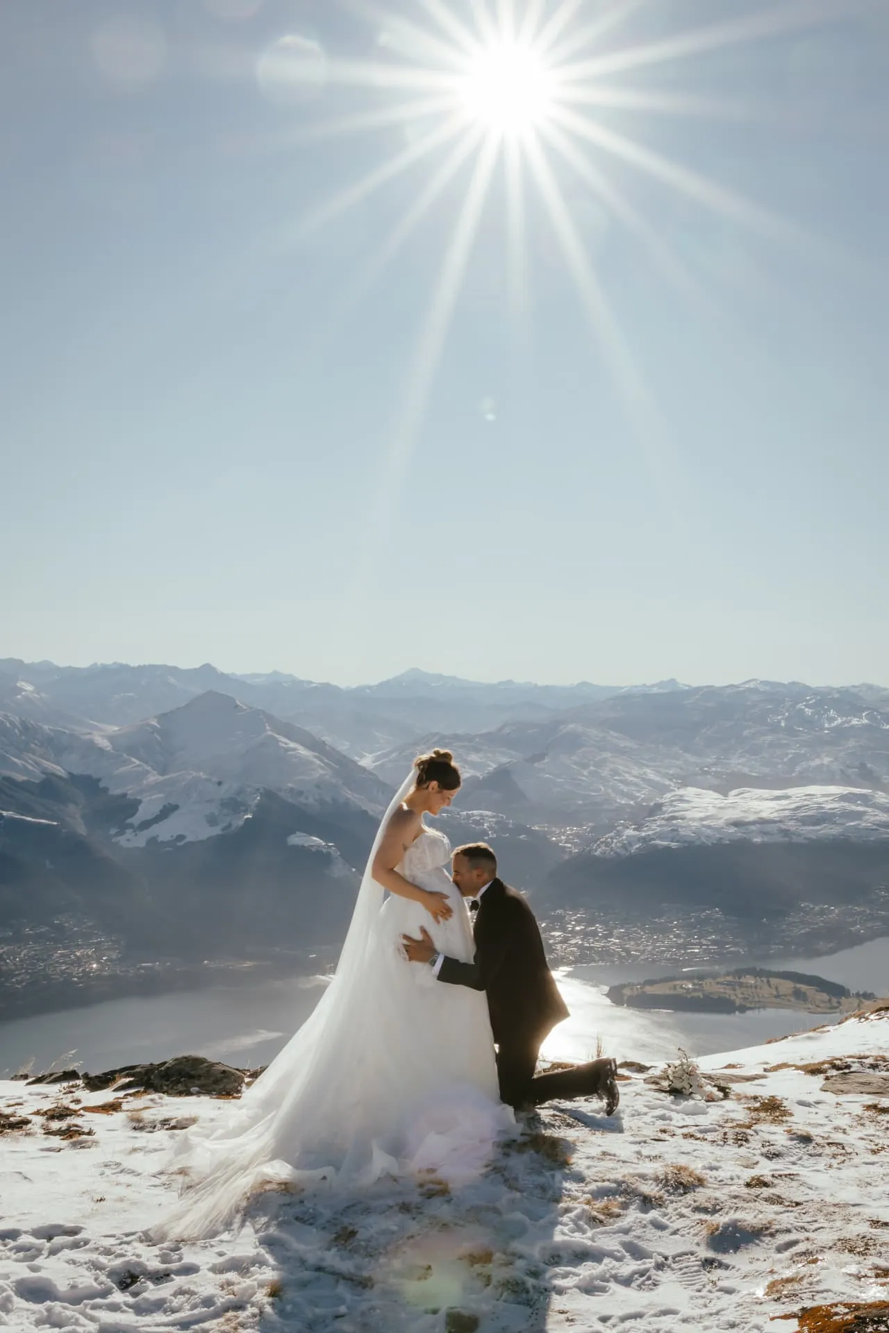 Groom kneeling before his bride in the snow on Cecil Peak under a bright winter sun.