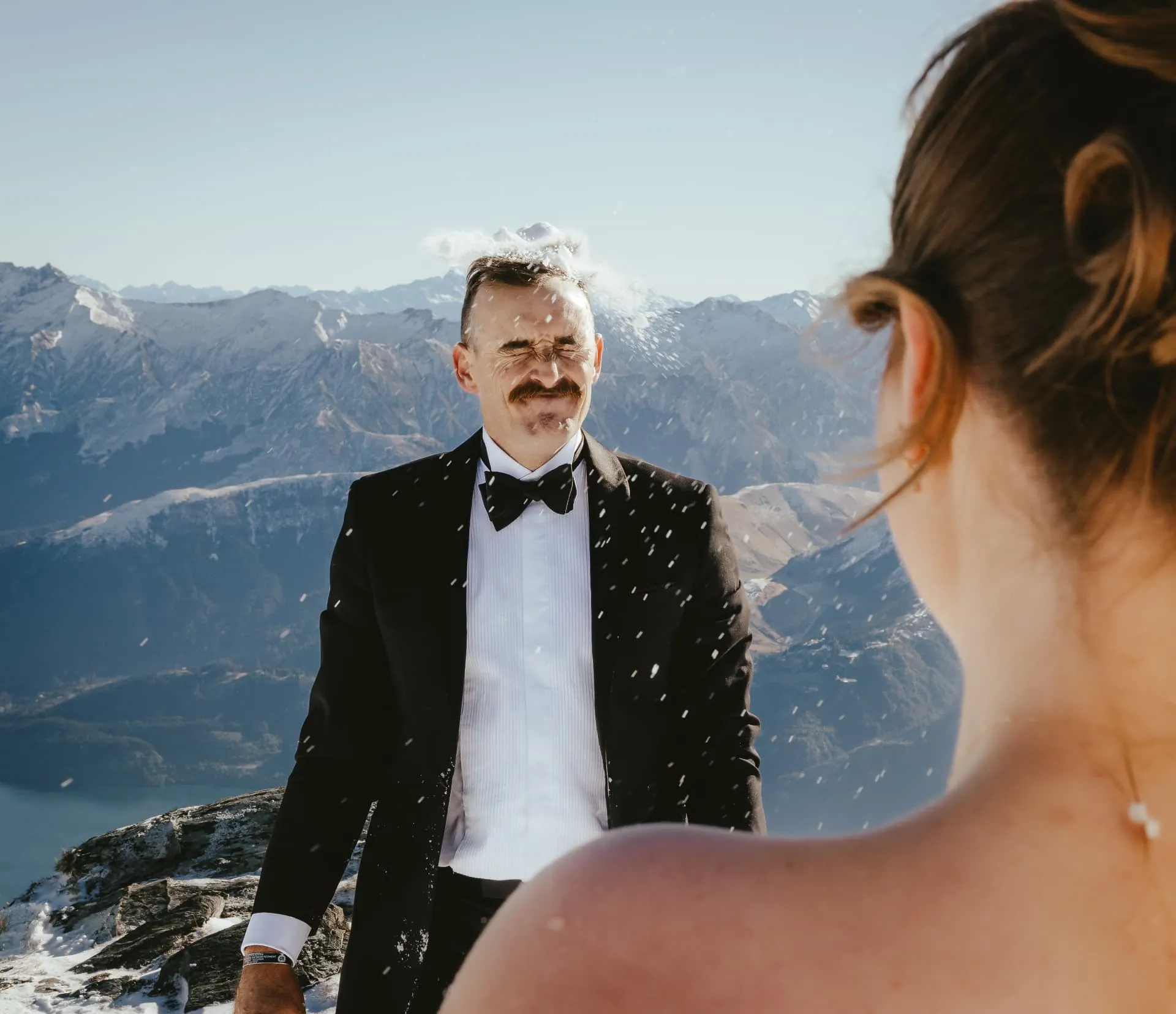 Groom bracing for a snowball over the bride\u2019s shoulder, snowy mountains behind, Cecil Peak.