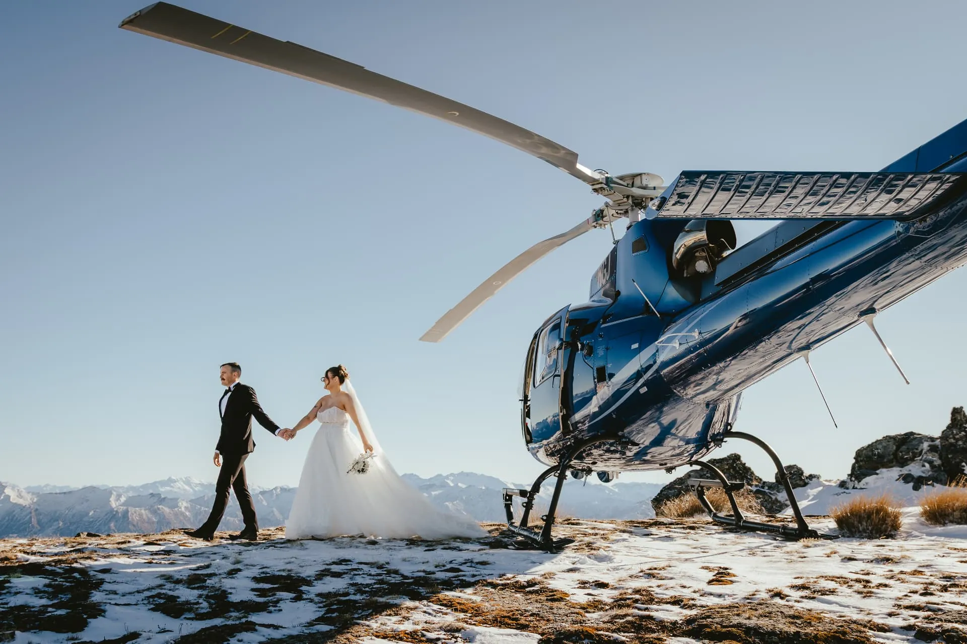 Couple walking hand in hand from the helicopter across snow on the Spur at Cecil Peak, end of winter.