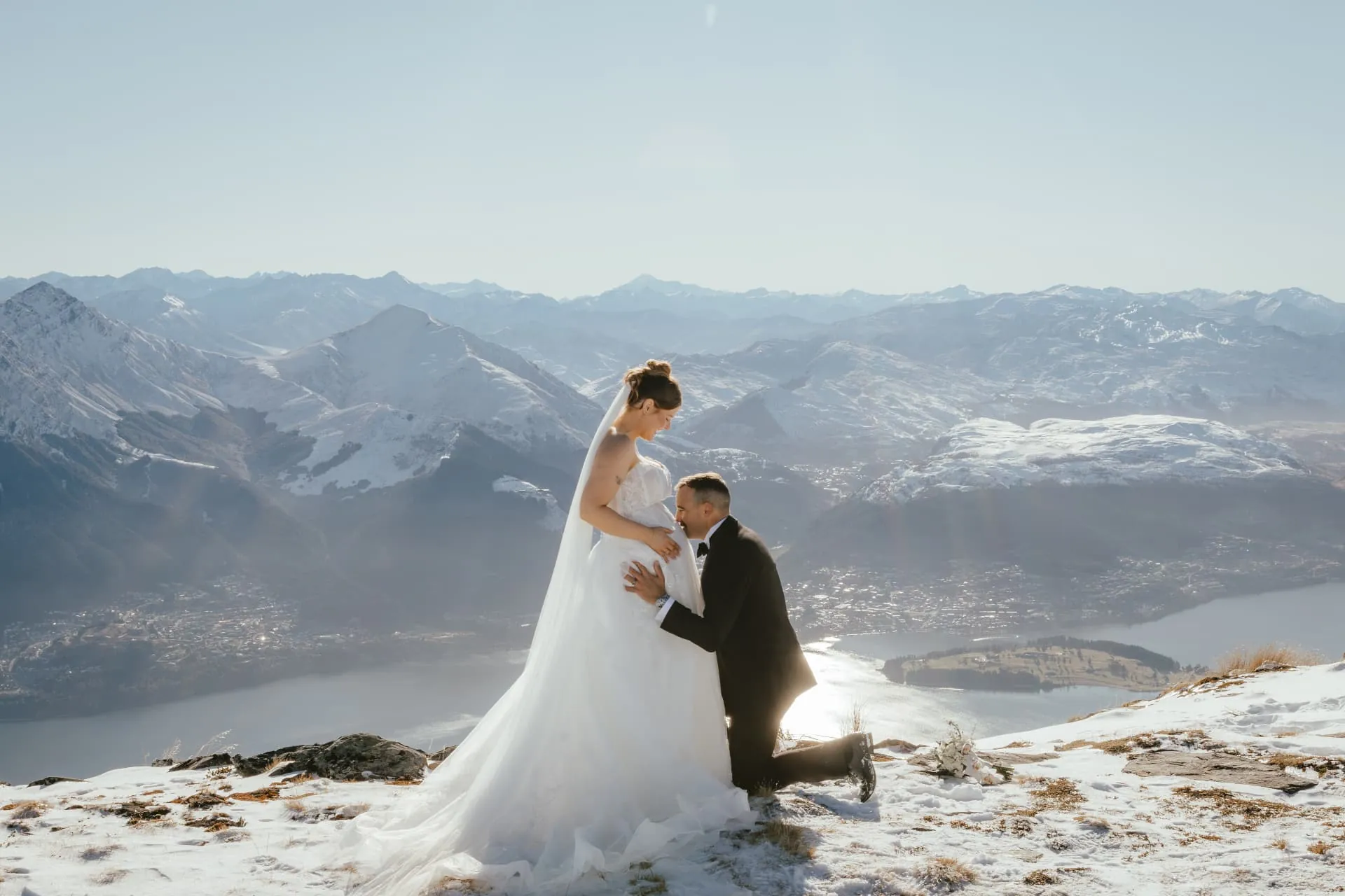 Groom kneeling in front of his bride on the snowy Spur at Cecil Peak, Queenstown and the lake beyond.