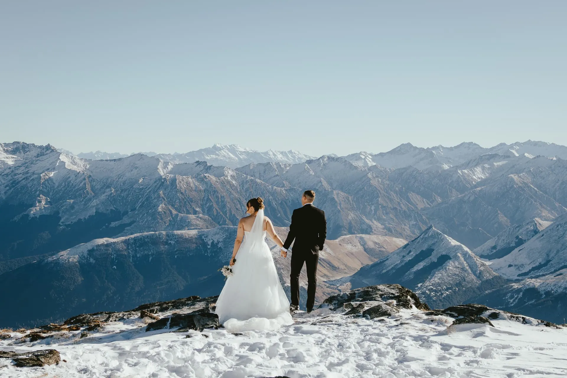 Couple in the snow on the Spur at Cecil Peak, Queenstown, with snowy mountains behind.