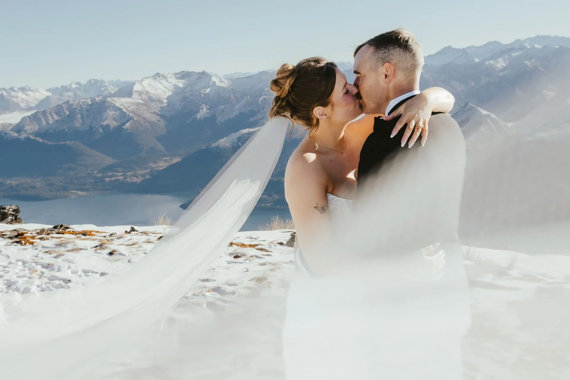 Couple kissing in the snow on Cecil Peak, the bride\u2019s long veil caught by the wind, snowy ranges behind.