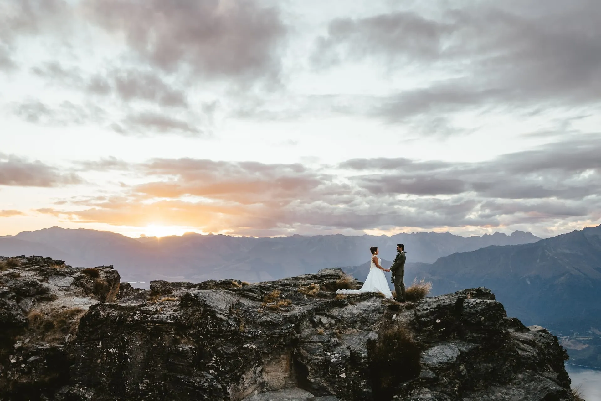 Wide view of a couple on the summit rock at Cecil Peak under a sunset sky, mountains in every direction.