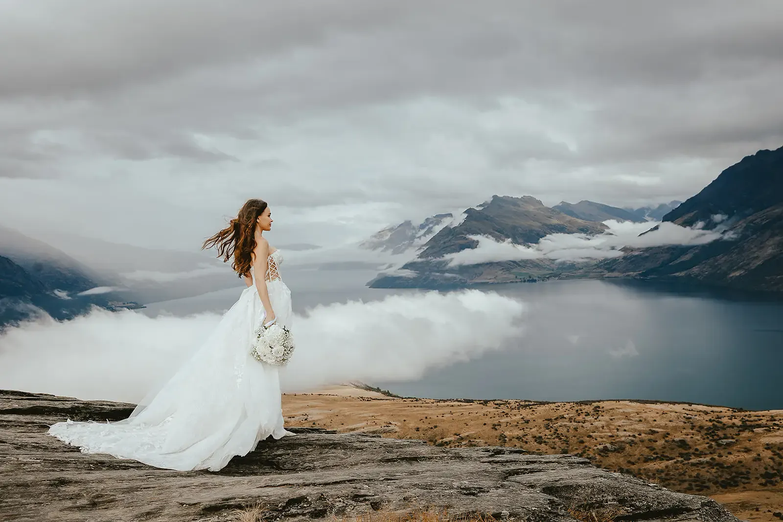 Bride on the western rock ledge at Deer Park Heights, hair in the wind, low cloud over Lake Whakatipu
