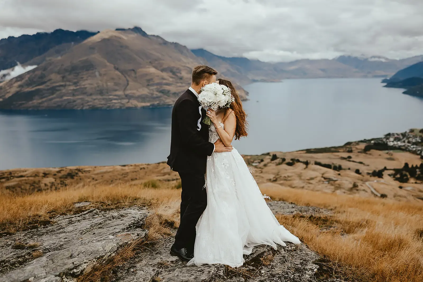 Couple kissing behind a white bouquet on a schist outcrop at Deer Park Heights, Lake Whakatipu behind