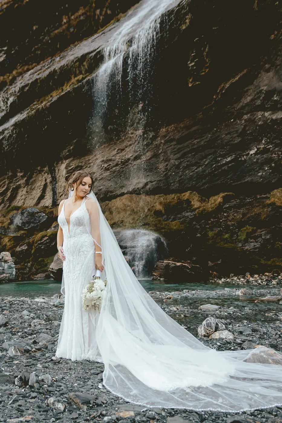 Bride with a long cathedral veil on the river stones below the main Earnslaw Burn waterfall in early winter