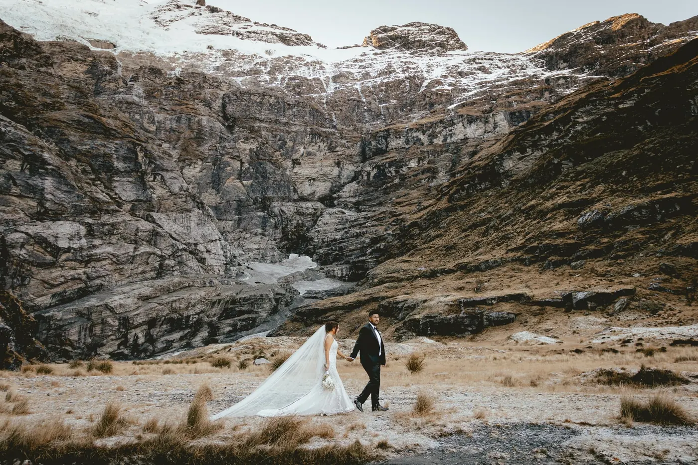 Bride and groom walking hand in hand across frosty tussock beneath the snow-dusted Earnslaw Burn glacier wall in early winter