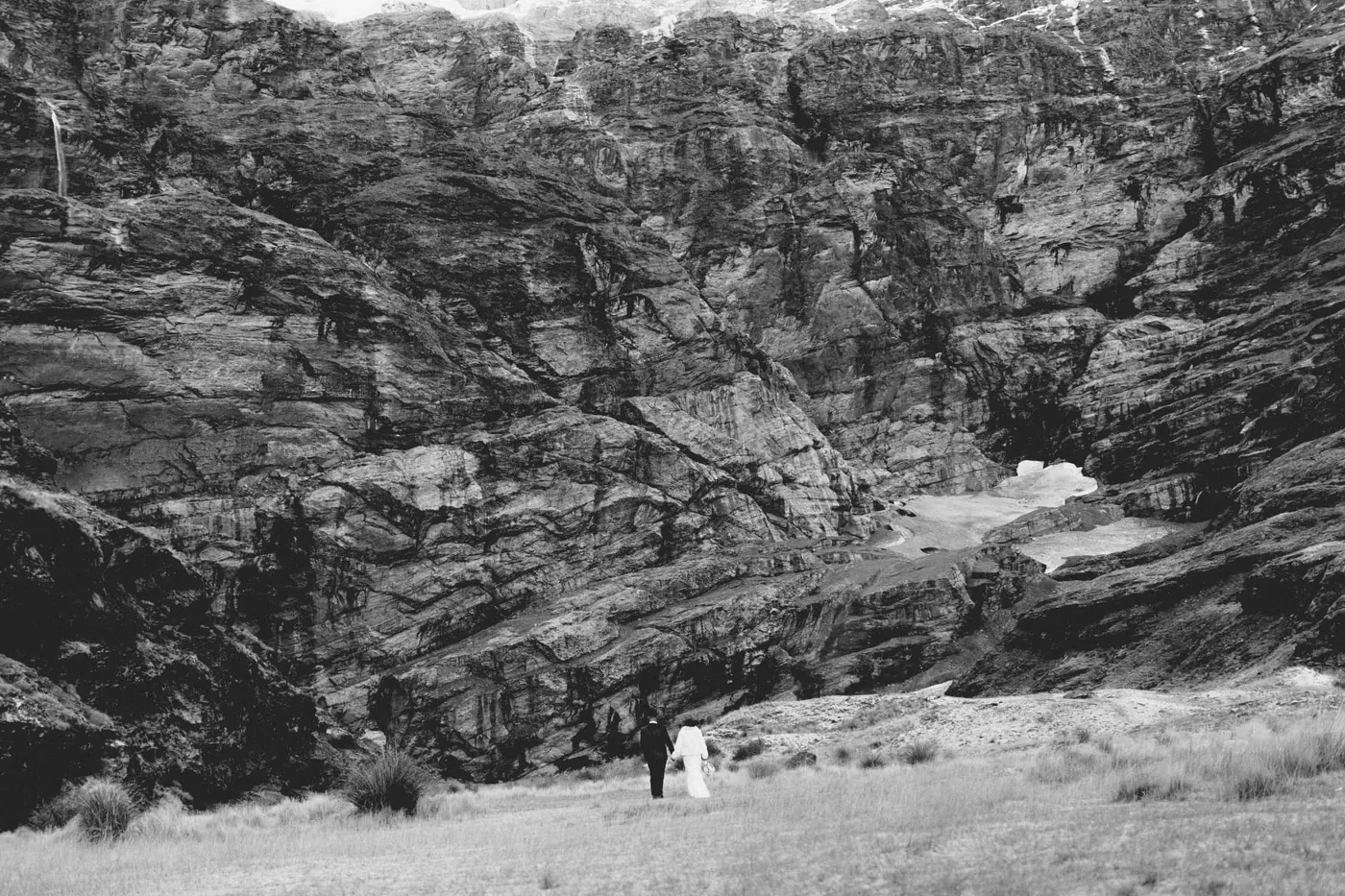 Black and white photo of a couple walking hand in hand, tiny beneath the Earnslaw Burn glacier wall in early winter