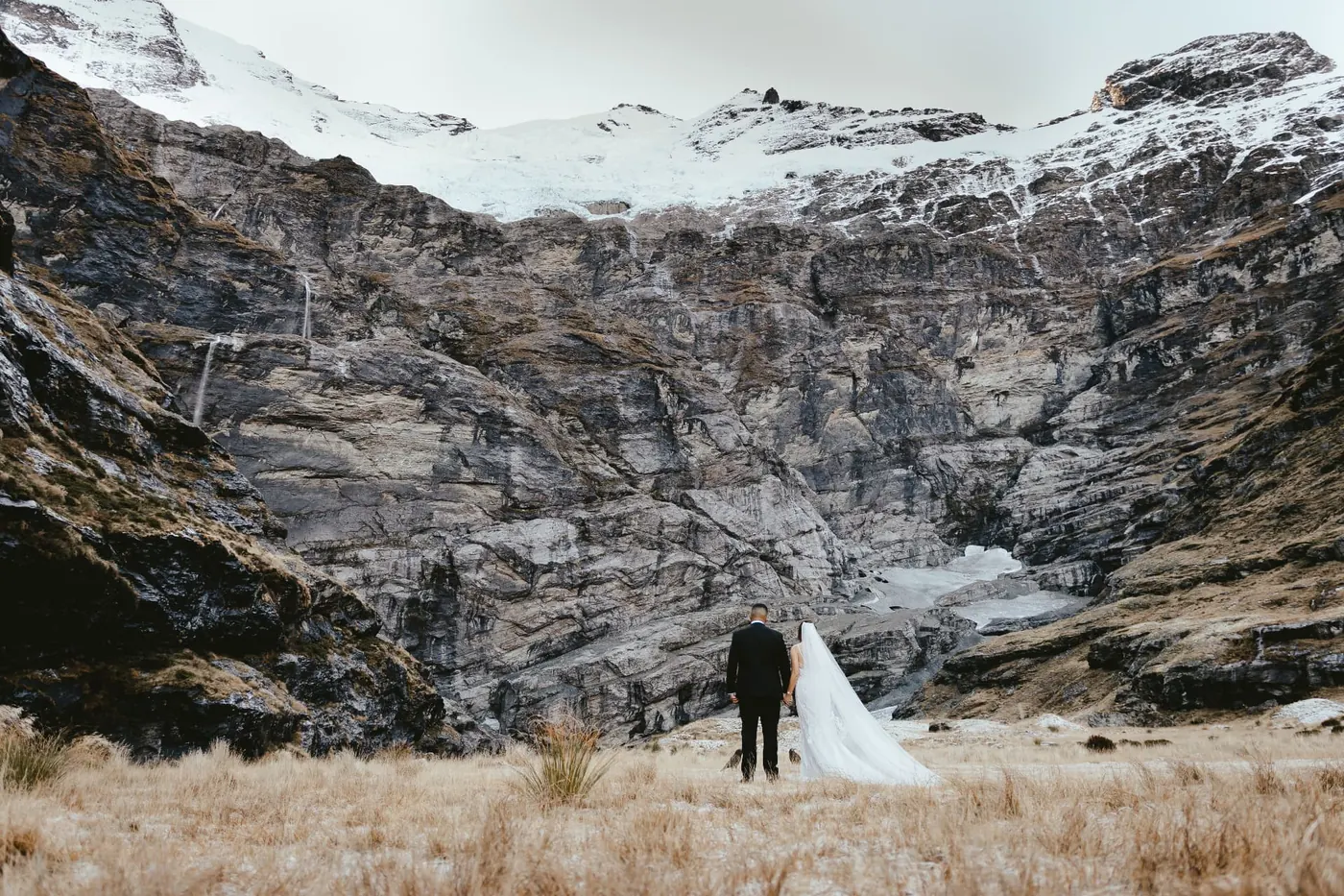Bride and groom standing in golden tussock facing the snow-capped Earnslaw Burn glacier wall at the beginning of winter