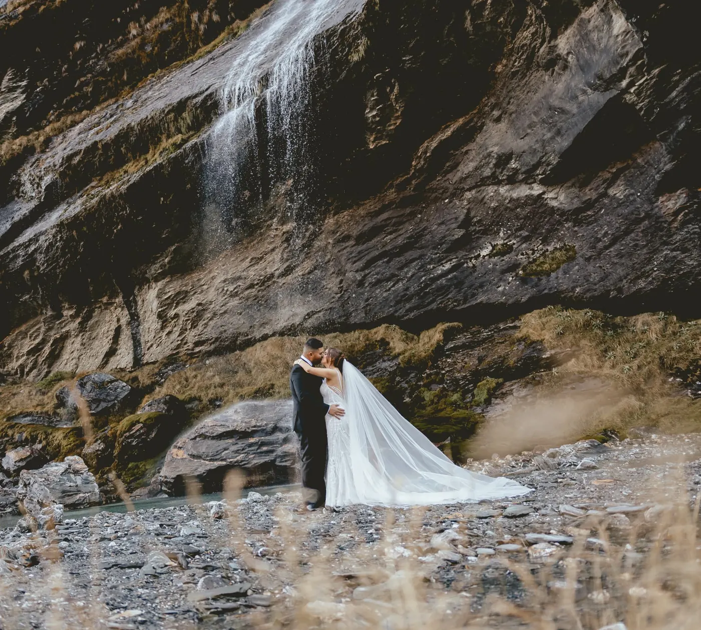 Couple embracing on the river stones beneath the main Earnslaw Burn waterfall during an early winter heli elopement