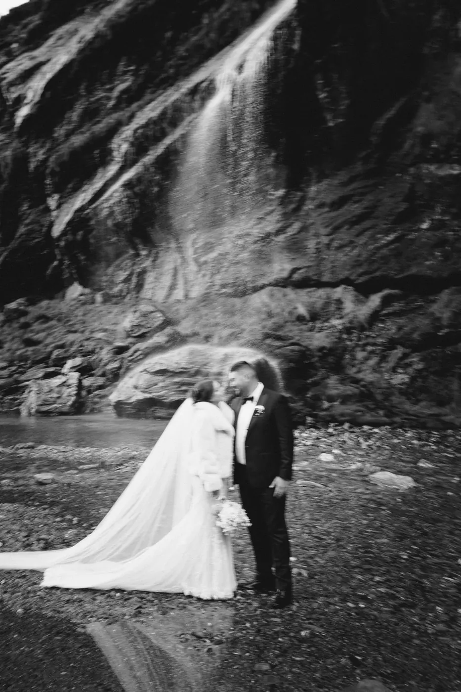 Black and white photo of a bride in a fur wrap kissing the groom beneath the main Earnslaw Burn waterfall