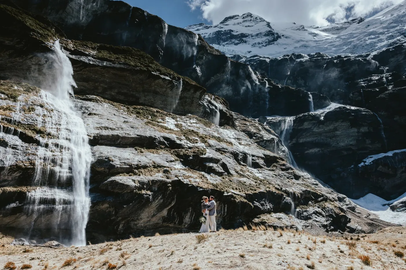Couple standing beneath the Earnslaw Burn waterfalls during their heli elopement in a glacier valley near Glenorchy.